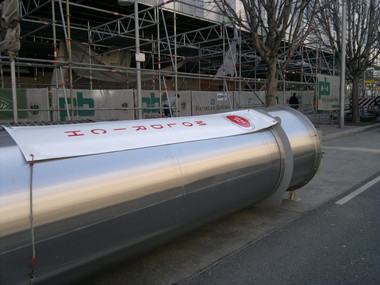 A large, shiny metal cylinder lies on its side on a street beside a construction site with scaffolding and bare trees in the background.