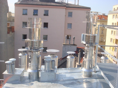 Rooftop with multiple metal ventilation pipes and chimneys, with pastel apartment buildings in the background.