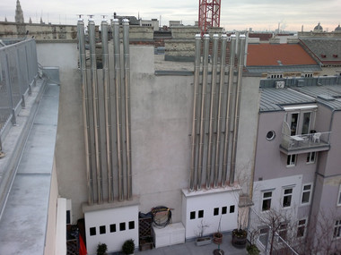 Rooftop view featuring tall vertical metal ventilation stacks along a white wall with a cityscape of rooftops in the background.