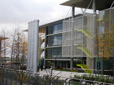 A modern glass-fronted building with a perforated metal facade and a tree-lined walkway in autumn, with people walking nearby.