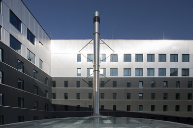 Modern office complex with many windows and a central glass-roofed area topped by a tall metal antenna against a blue sky.