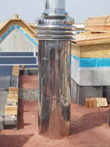 A tall, reflective metal roof vent pipe on a construction-site roof with wooden framing, bricks, and blue-trimmed roofing in the background.