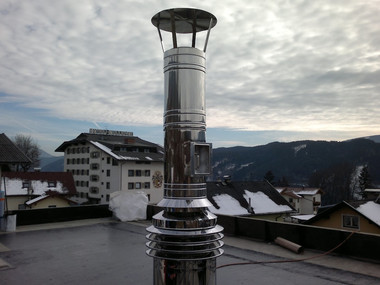 Shiny metal chimney vent on a flat roof with houses and mountains in the background.