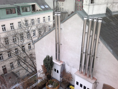 Rooftop terrace with tall metal vent pipes along a white wall, a small tree and potted plants, and neighboring apartment buildings.