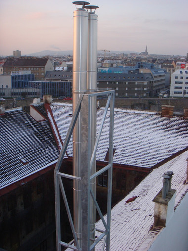 A tall metal exhaust stack with a supporting framework rises from a snow-covered rooftop overlooking a city skyline.