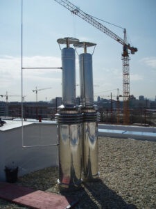 Rooftop scene featuring two tall silver vent pipes with a reflective base and a construction crane in the background.