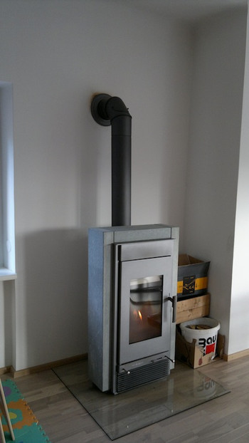 A silver wood-burning stove with a vertical black flue pipe in the corner of a bright room, with storage boxes beside it.