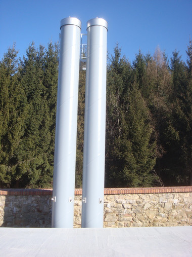 Two tall gray metal pipes stand side by side in front of a stone wall with trees and a blue sky in the background.