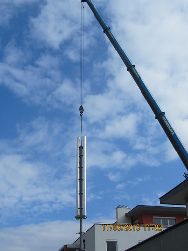 A construction crane hoists a tall vertical metal pole beside a building against a blue sky.