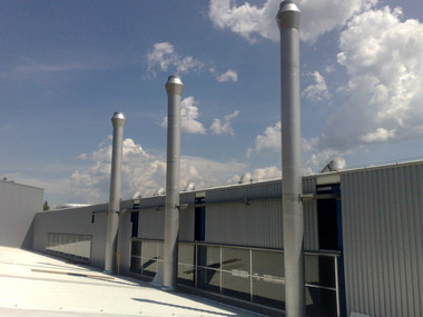 Rooftop of an industrial building with four tall metal vent stacks and railing, under a blue sky with clouds.