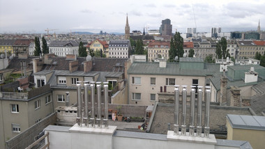 Rooftop view of a dense urban landscape with several metal chimneys in the foreground and a distant church spire under a cloudy sky.