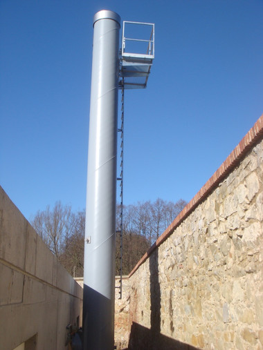 Tall gray metal ventilation stack with a small platform and ladder at the top, standing between stone walls under a clear blue sky.