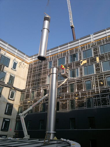 A crane lifts a tall stainless steel ventilation duct into place on the exterior of a partially constructed building, with scaffolding and a worker in a lift nearby.