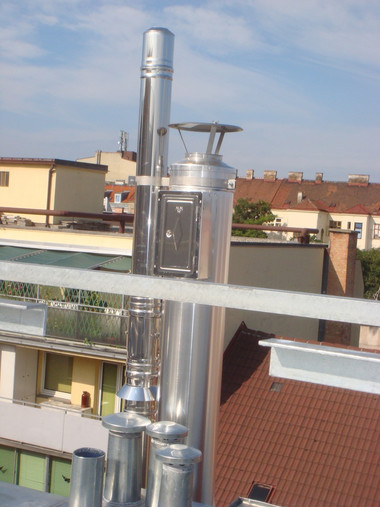 Tall stainless steel chimney stack rising from a rooftop with residential buildings and blue sky in the background.