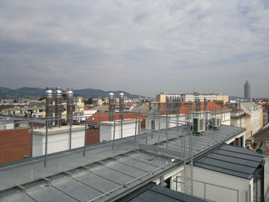 Rooftop view over a city with red-tiled roofs, a distant tall building, and hills on the horizon under a cloudy sky.