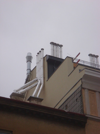 Rooftop of a beige building with multiple chimneys and vent pipes against a cloudy sky.