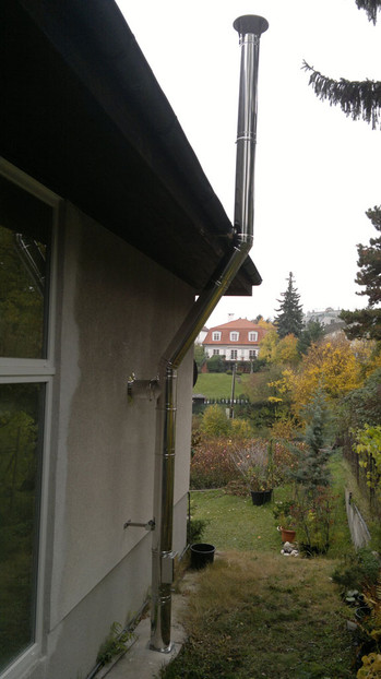 Vertical metal chimney attached to the exterior wall of a house, with a garden and a distant red-roofed house among trees.