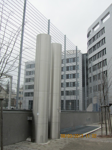 Two tall stainless steel ventilation ducts rise in a paved courtyard beside a modern grey office building, with a chain-link fence and bare trees.