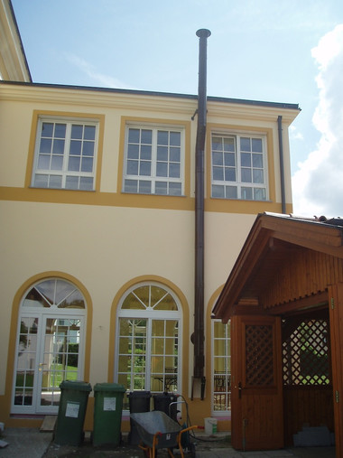 Two-story yellow building with arched ground-floor windows, rectangular upper windows, a tall black downspout along the wall, and a wooden gazebo to the right.