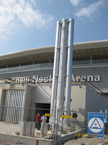 A construction site in front of a modern arena with tall vertical metal pipes and a blue safety sign.