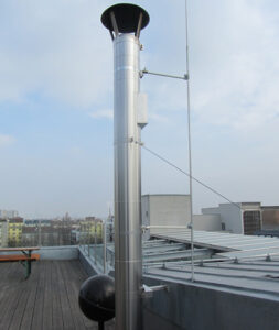 Tall stainless steel rooftop ventilation pipe with a black cap and supporting cables on a wooden deck, with a city skyline in the distance.
