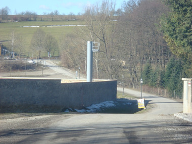 Rural road curving past a low stone wall with a tall metal sign post, bare trees, and patches of snow on the ground.