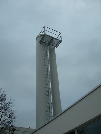 Tall tower with an external ladder and a top observation platform against a cloudy sky.