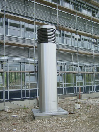 A tall silver cylindrical vent with a slatted top sits on a concrete base in front of a multi-story glass-and-steel building.