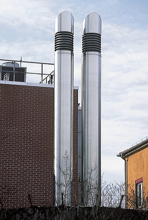 Two tall silver ventilation stacks on a rooftop beside a brick building against a blue sky.