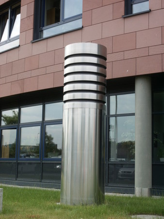 Tall stainless steel cylindrical sculpture with black horizontal bands stands on a grassy patch beside a brick building.