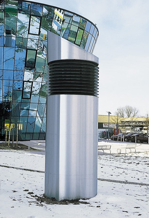 A tall silver cylindrical vent-like sculpture with black horizontal slats stands on snow-covered ground beside a curved glass building.