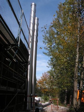 Construction site with tall metal chimneys and scaffolding on the left and autumn trees on the right under a blue sky.