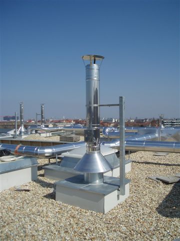 On a gravel rooftop, a tall metal ventilation chimney rises beside other rooftop equipment against a clear blue sky.