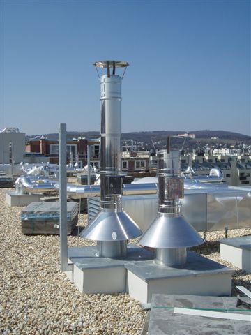 Rooftop with metal ventilation stacks on a gravel surface and a distant city skyline under a clear blue sky.