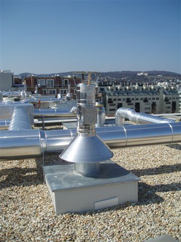 Metal rooftop ventilation pipes and a vent on a gravel-covered roof with a city skyline in the distance.
