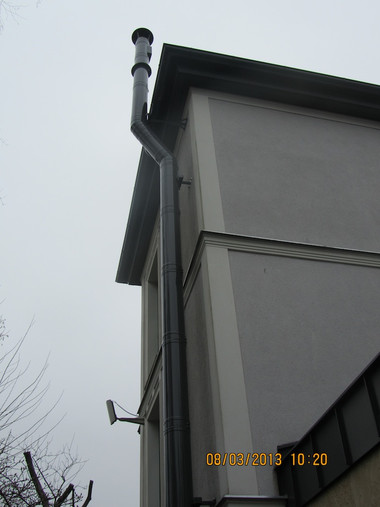 Corner of a gray building with a vertical downspout and gutter along the roof, against an overcast sky.