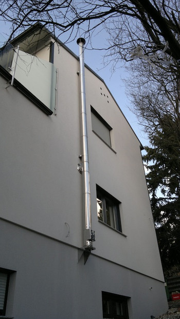 Side view of a white multi-story building with a tall vertical pipe along its exterior, with windows and trees against a blue sky.