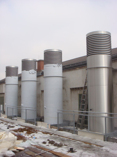 Row of tall cylindrical metal ventilation stacks on a rooftop with a metal railing and a ladder.