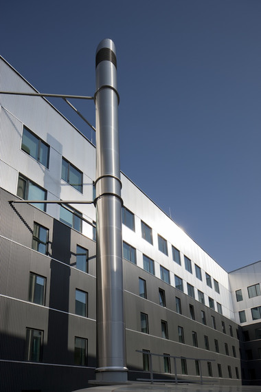 A modern office building with a tall cylindrical metal vent in the foreground against a clear blue sky.
