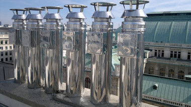 A row of tall, shiny metal vent pipes on a rooftop with a building and blue sky in the background.