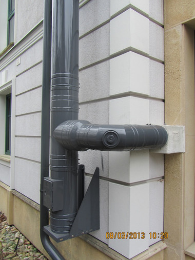 Gray metal rain downspout with a short horizontal outlet mounted on a light-colored exterior wall.