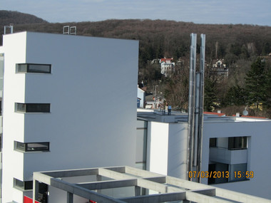 White modern apartment buildings with a metal rooftop framework, set against a hilly, tree-covered background.
