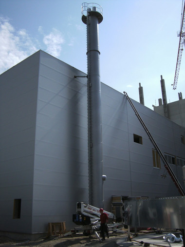 Industrial facility with a tall vertical exhaust stack beside a gray building, with construction equipment and a crane visible in the background.