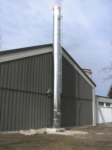 A tall silver vent stack with a ladder running up its side stands beside a dark metal building.