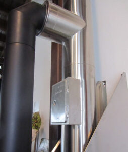 A small metallic electrical junction box mounted on a vertical shiny pipe with an elbow joint, next to a black insulated pipe.