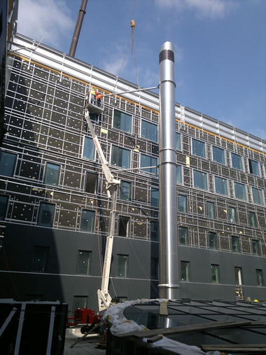 Exterior of a multi-story building under renovation with scaffolding on the facade, a boom lift, and a tall metal vent pipe against a blue sky.