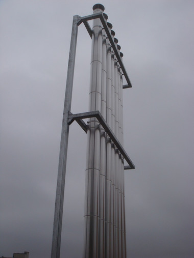 Tall metal industrial structure with vertical pipes and walkways against a cloudy sky.
