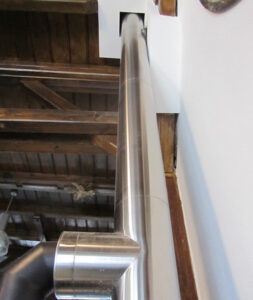 Shiny stainless steel HVAC ducting with an elbow joint runs along a white wall in an unfinished attic with wooden ceiling beams.