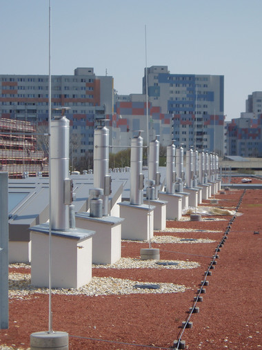 Row of white metal ventilation shafts on a red gravel rooftop with apartment buildings in the background.