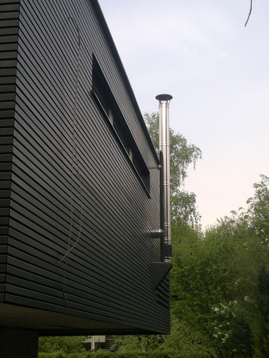 Dark, horizontally slatted exterior wall of a modern building with a tall metal vent pipe and green trees in the background.